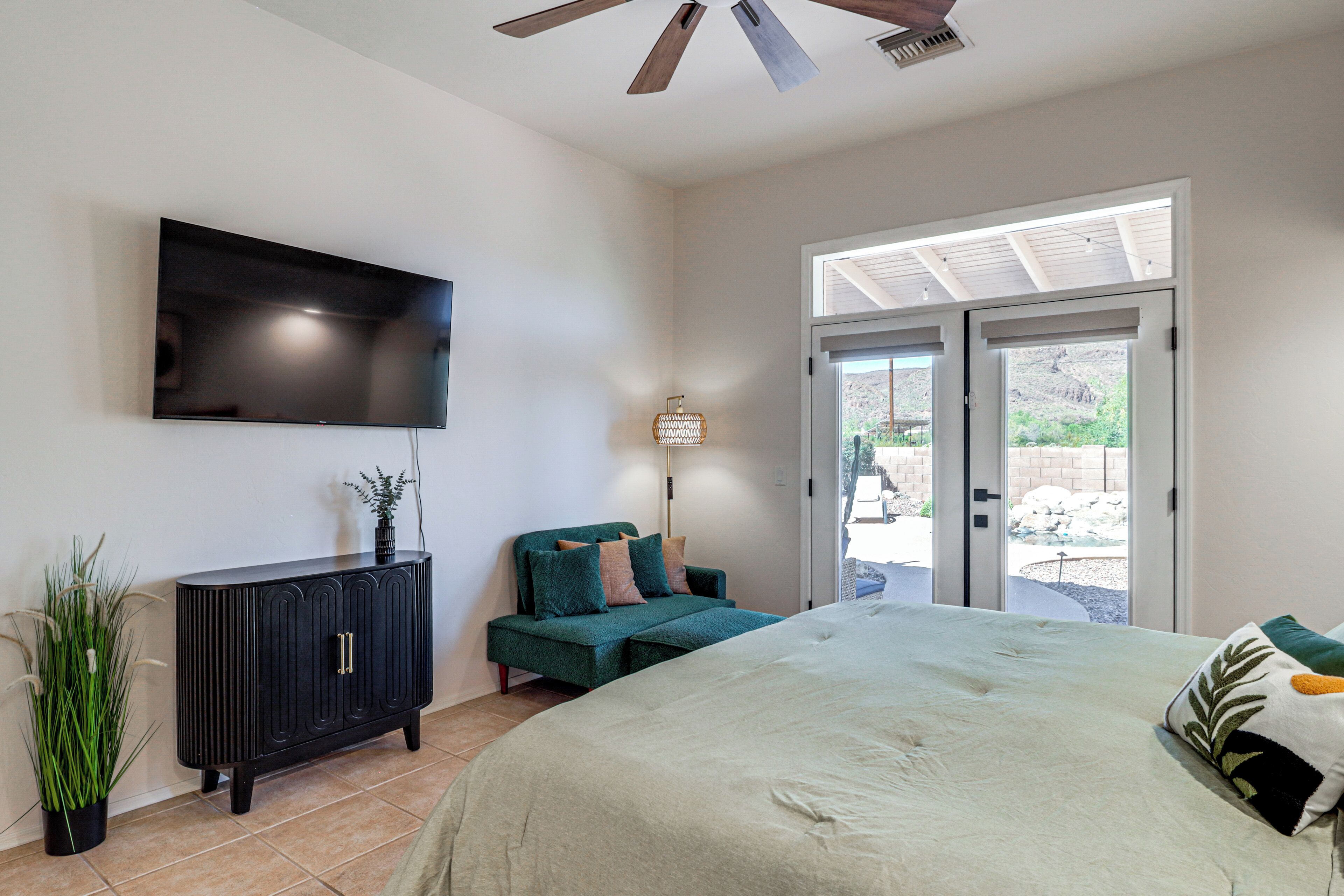 A bedroom with a bed, a TV mounted on the wall, a dark-colored cabinet, a green sofa, and a sliding glass door leading to an outdoor area.
