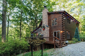 A rustic cabin with a stone chimney, wooden deck, and a sign indicating it's a Christmas cabin in Mineral Bluff, GA. - The Christmas Cabin | Creekside Hot Tub Escape (Mineral Bluff)