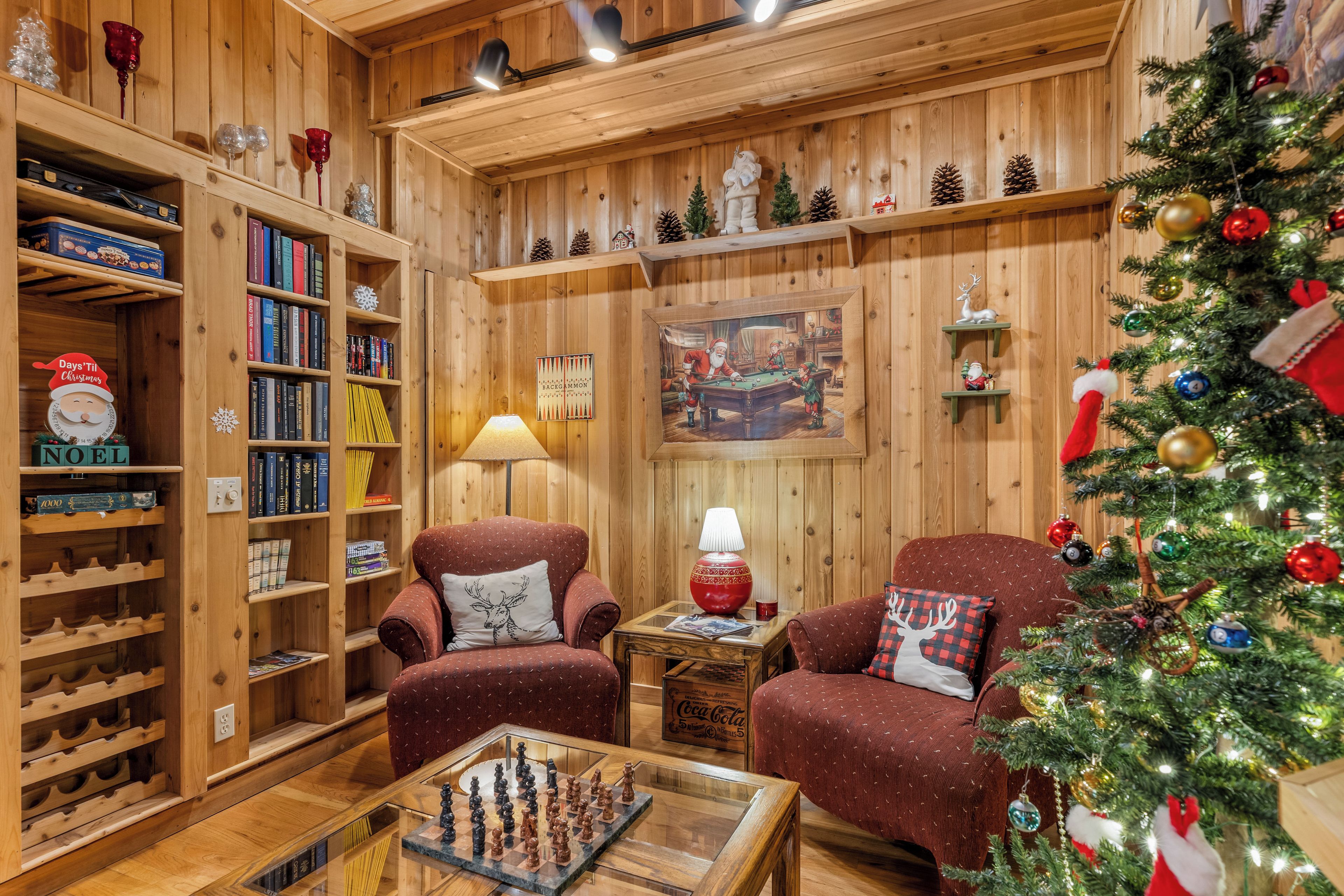 A cozy room with wooden walls, a Christmas tree, two armchairs, a coffee table with a chess set, and shelves with books and decorative items.