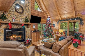 Interior acollidor d'un xalet de fusta amb una llar de pedra, un arbre de Nadal, sofàs de pell i una televisió penjada a la paret.