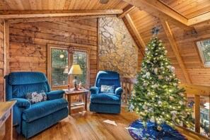A cozy room with wooden walls, a stone accent wall, two blue armchairs, a Christmas tree, and a window with a view of trees. - The Christmas Cabin | Creekside Hot Tub Escape (Mineral Bluff)