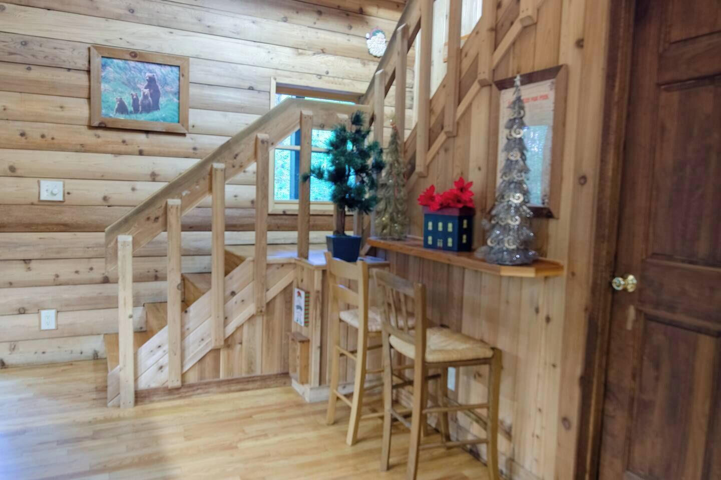 A wooden interior with a staircase, a small Christmas tree, a shelf with decorations, and a framed picture on the wall.