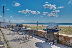 Eine Außenpatio mit Tischen, Stühlen, einem Grill und Blick auf Strand und Meer.