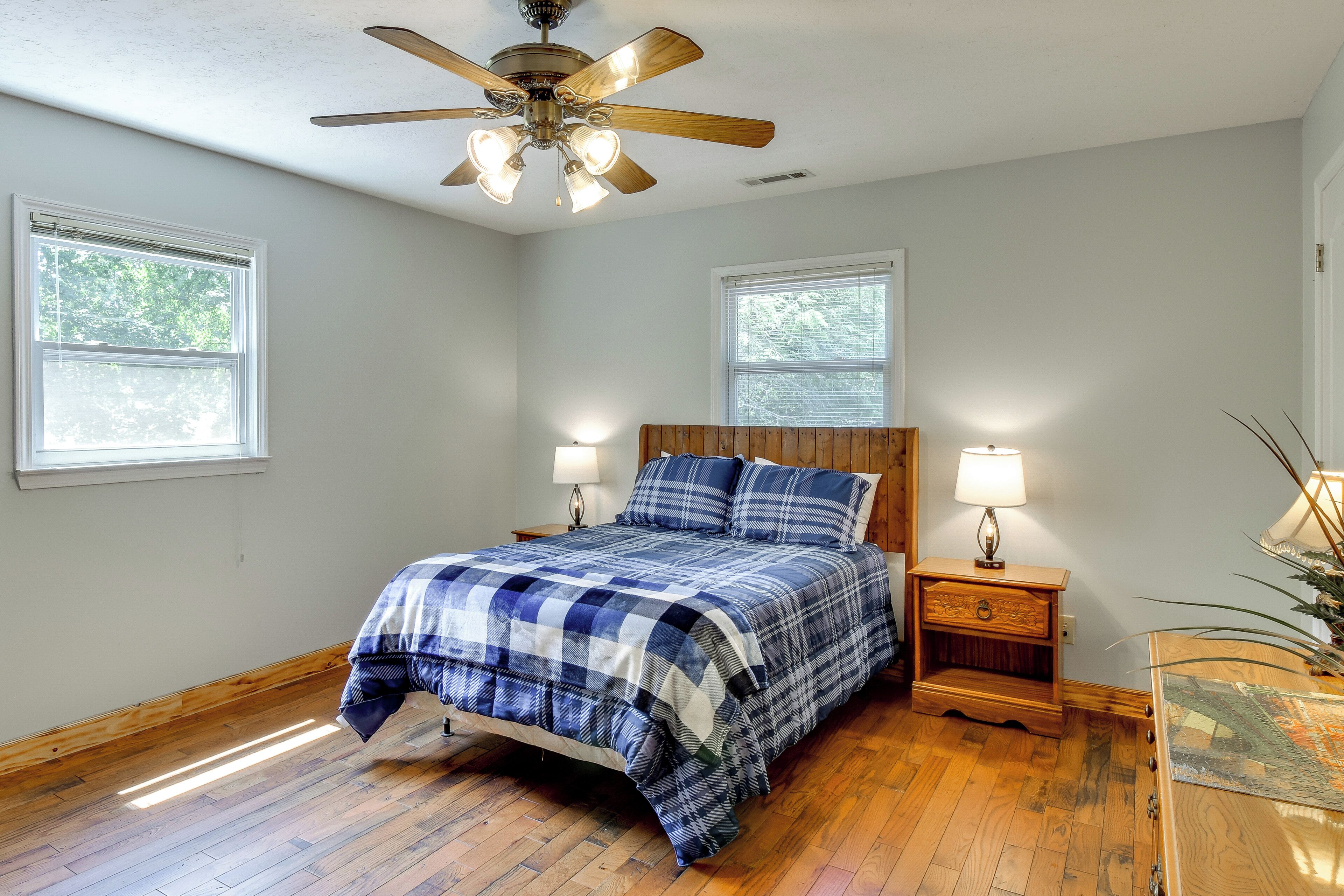 A bedroom with a wooden floor, a bed with a blue and white checkered bedspread, two bedside tables with lamps, and a ceiling fan.