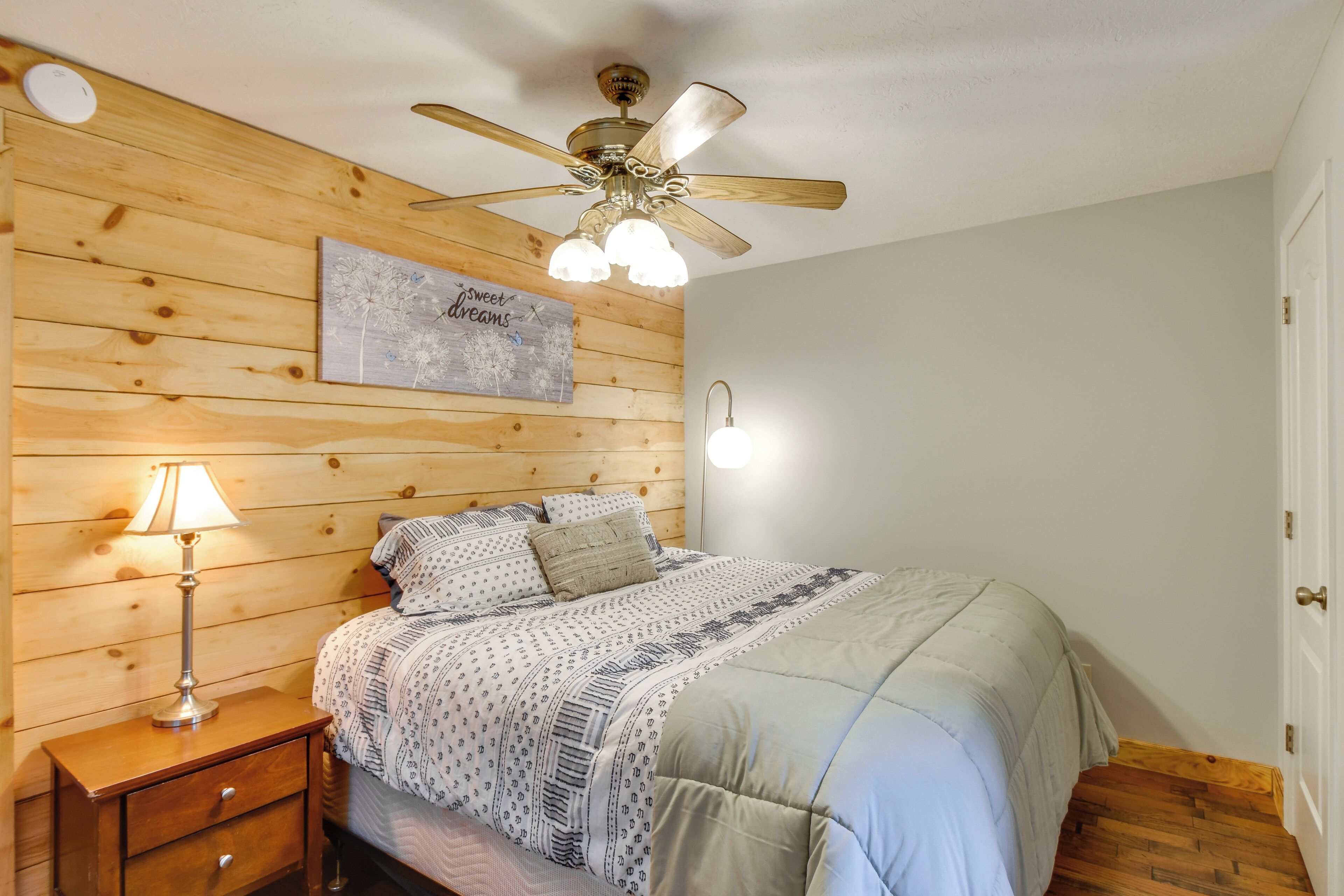 Quarto com cabeceira de madeira, ventilador de teto, luminária de cabeceira, cama com roupa de cama estampada e luminária fixada na parede.
