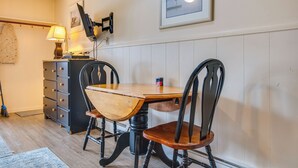 A dining area with a wooden table and chairs, a lamp, a television mounted on the wall, and a framed picture. - Steps to Beach! OOB Studio w/ Grill Access (Old Orchard Beach)