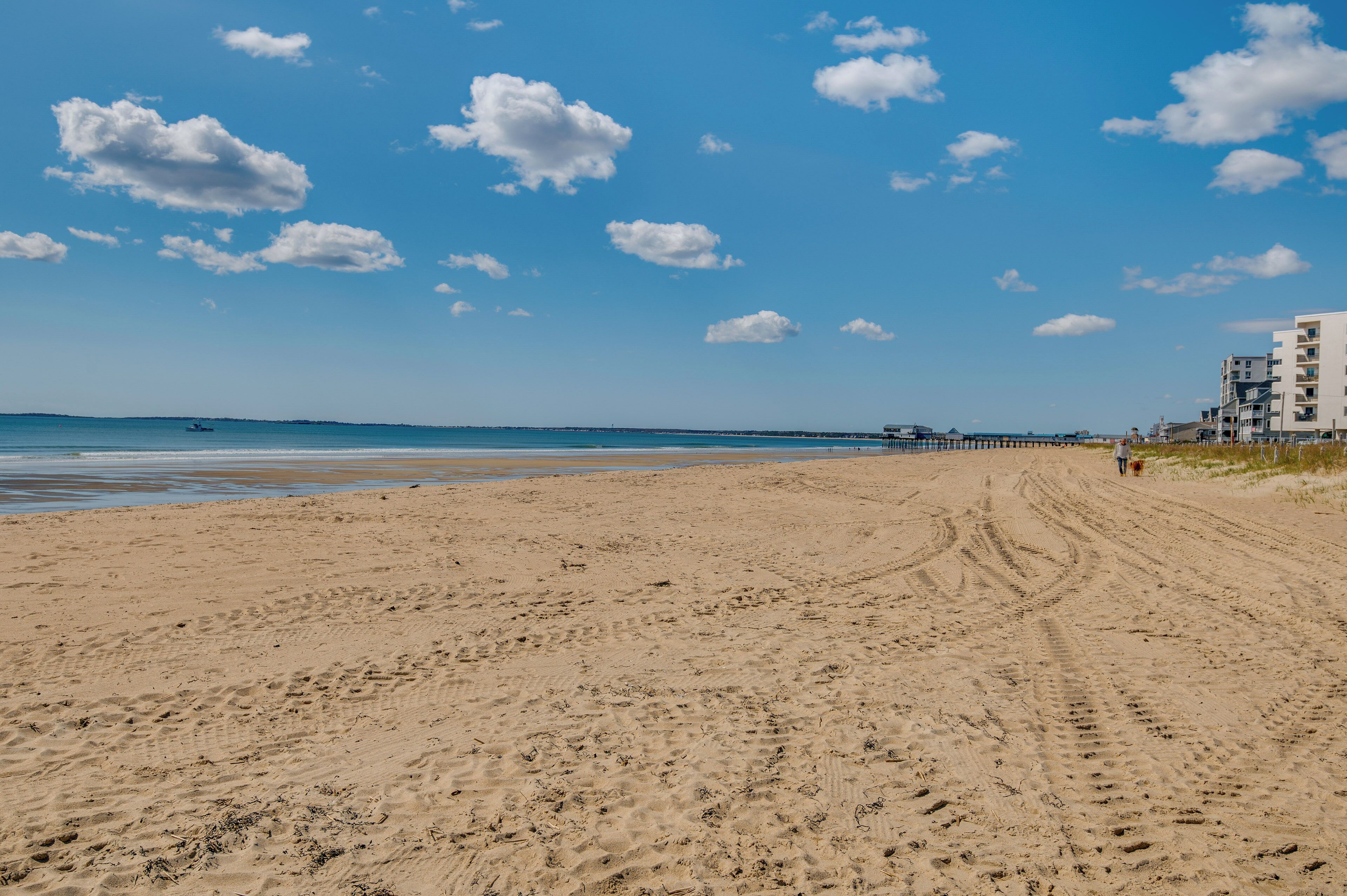 A sandy beach with footprints, a pier in the distance, and a building with multiple floors.
