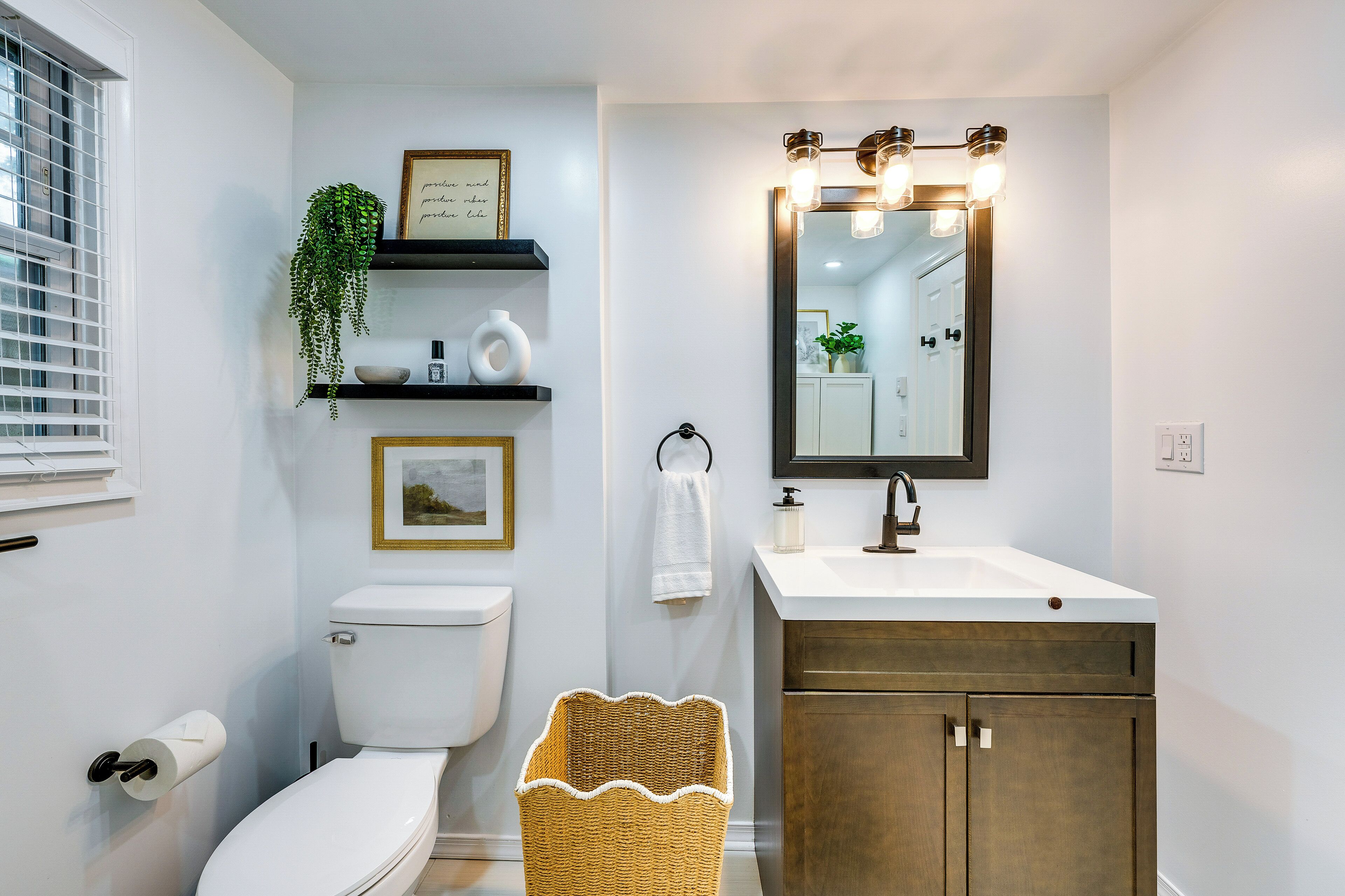 A bathroom with a toilet, sink, mirror, shelves with plants, and a wicker basket.