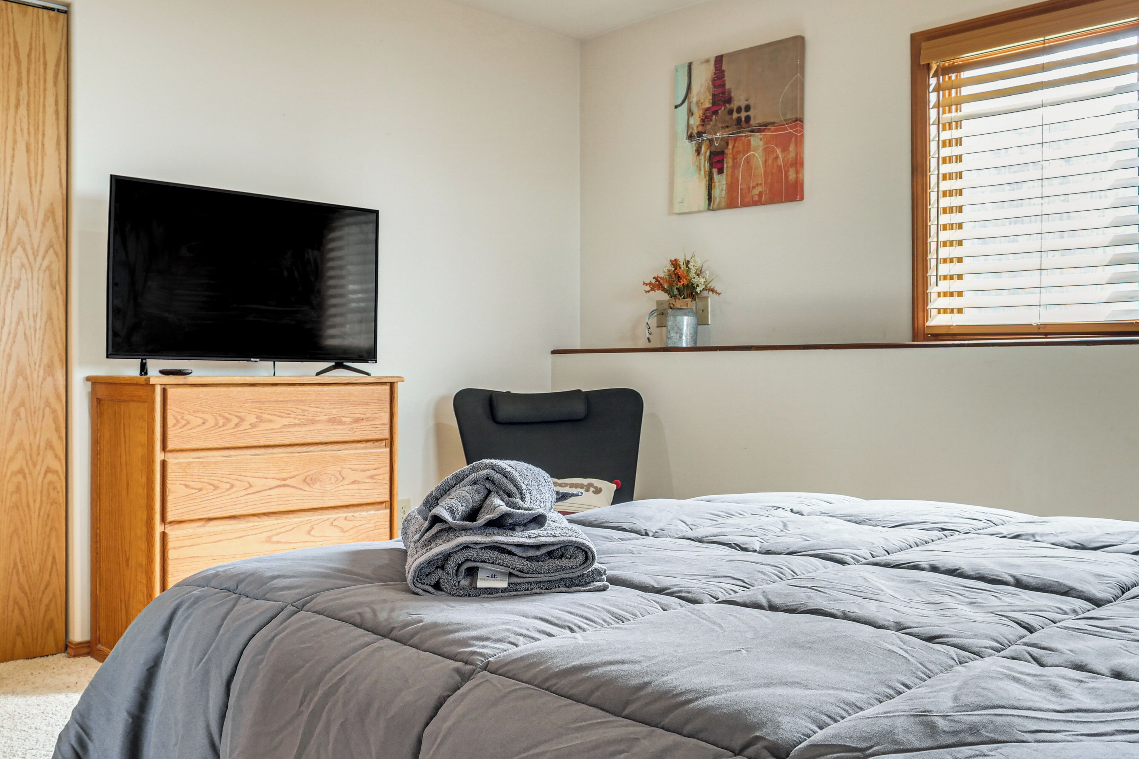 A bedroom with a bed, a television, a wooden dresser, a chair, and a window with blinds.