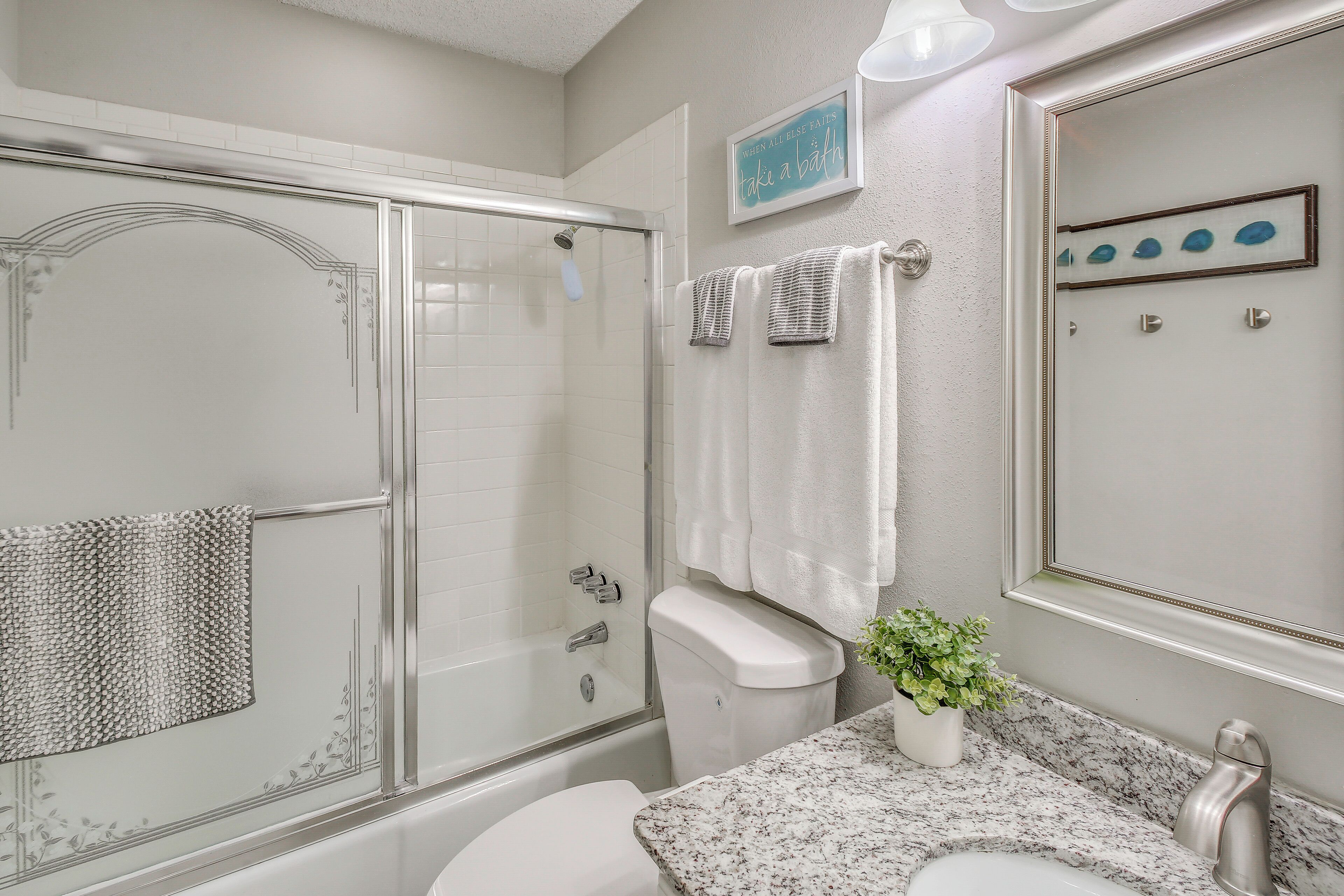 Un baño con cabina de ducha de vidrio, inodoro, lavamanos con encimera de mármol y una planta en maceta.