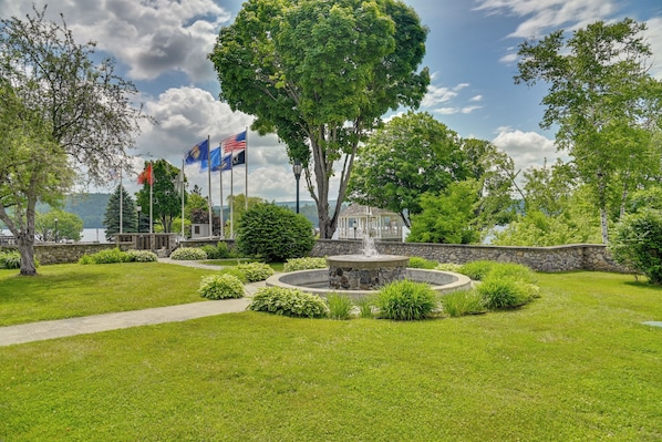 A landscaped garden with a fountain, flags, and a view of a body of water. - Walk to Schroon Lake! Studio w/ Shared Fire Pit (Schroon Lake)
