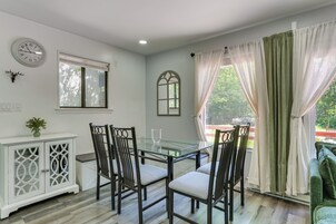 Un comedor con una mesa de vidrio, cuatro sillas, un armario blanco con detalles decorativos, un reloj y una ventana con persianas.