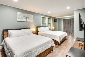 A hotel room with two beds, a painting, a window with blinds, a television, and a small table.