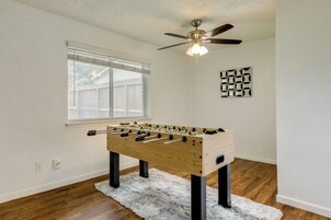 Una habitación con mesa de fútbol de madera, ventilador de techo, ventana con persianas y alfombra blanca.