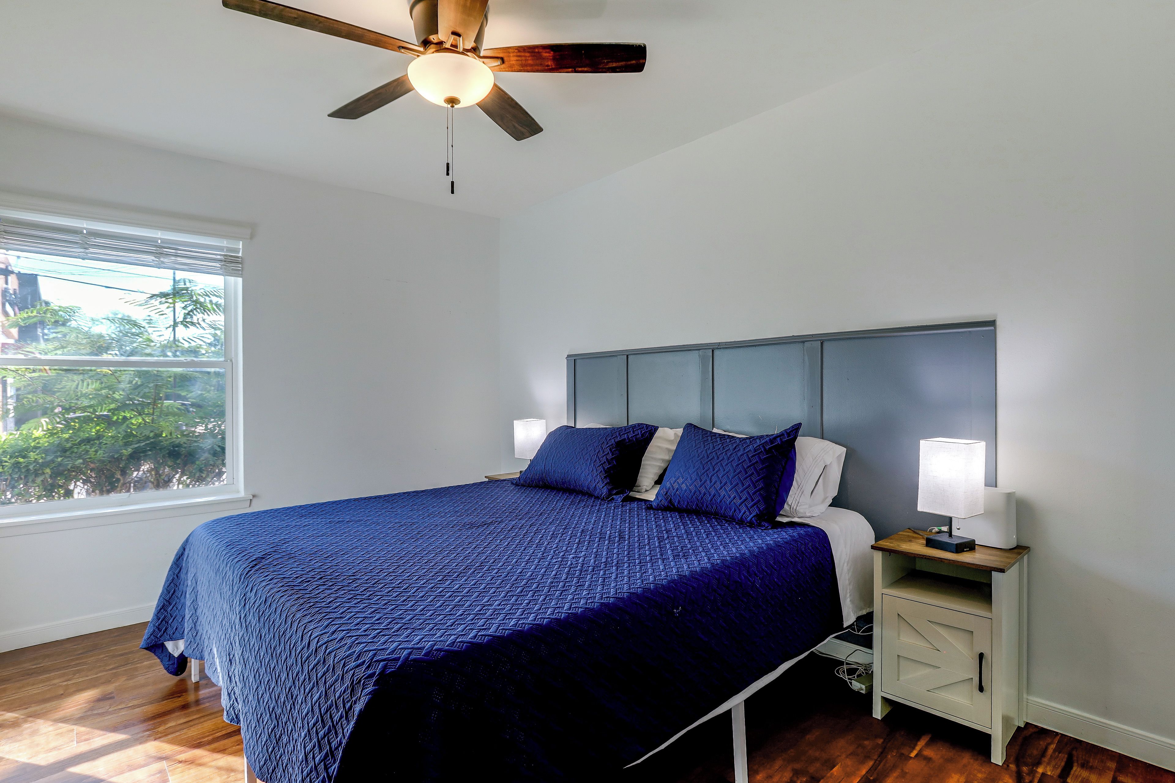 Une chambre à coucher comprenant un lit, une table de chevet, un ventilateur de plafond, une fenêtre et une vue sur la verdure à l’extérieur.