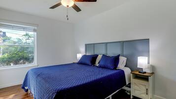 Une chambre à coucher comprenant un lit, une table de chevet, un ventilateur de plafond, une fenêtre et une vue sur la verdure à l’extérieur.