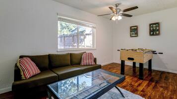 A living room with a glass coffee table, a sofa with striped pillows, a wooden foosball table, a ceiling fan, and a window with blinds.