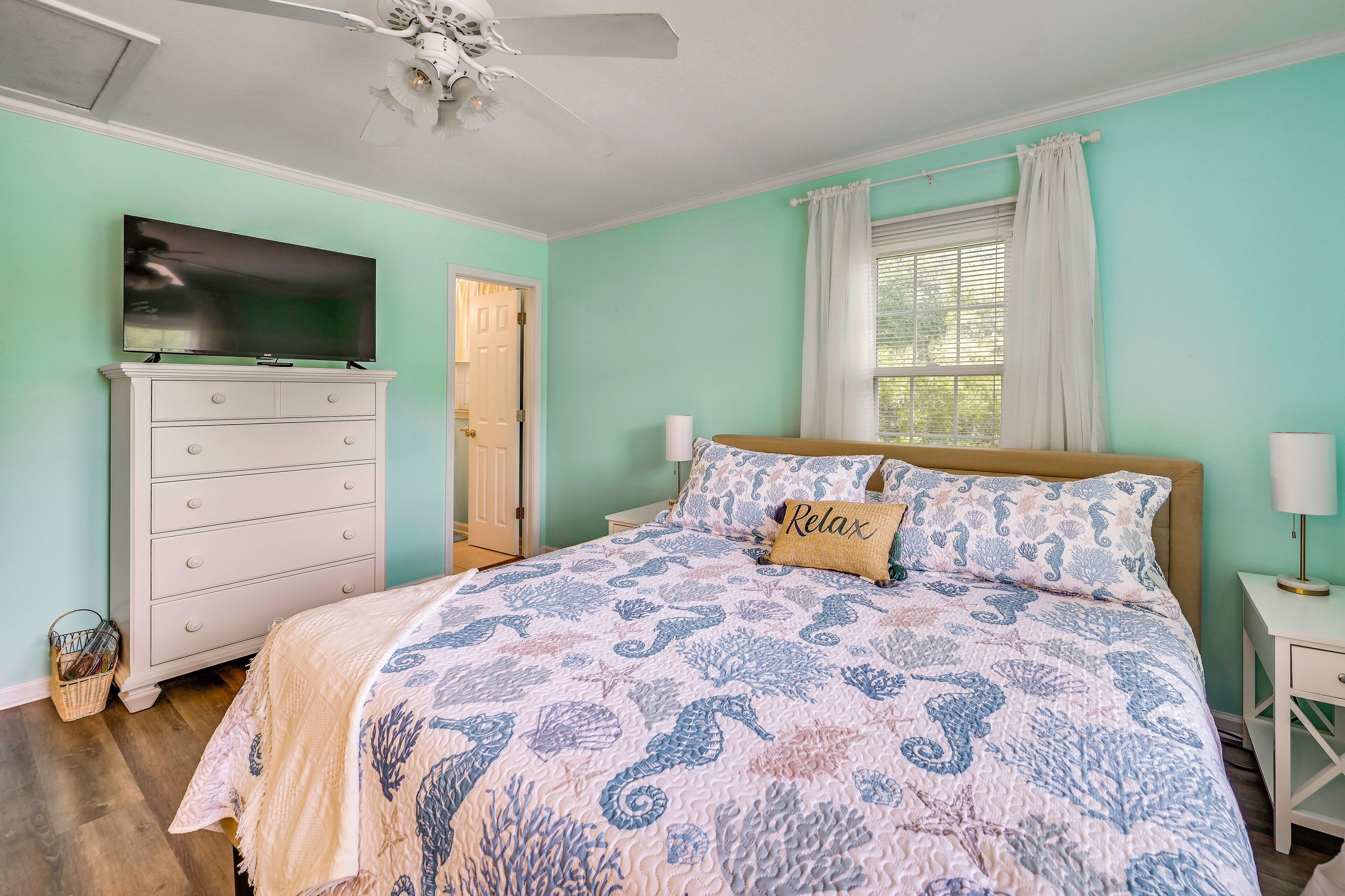 A bedroom with a bed, a dresser, a television, a ceiling fan, and a window with curtains.