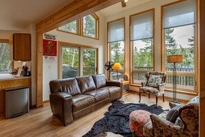 Ein gemütliches Wohnzimmer mit einem Ledersofa, einem gemusterten Sessel, einem Teppich und großen Fenstern mit Blick auf Bäume.