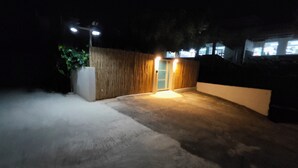 Un espacio exterior nocturno con suelo de concreto, cerco de madera y una puerta iluminada.