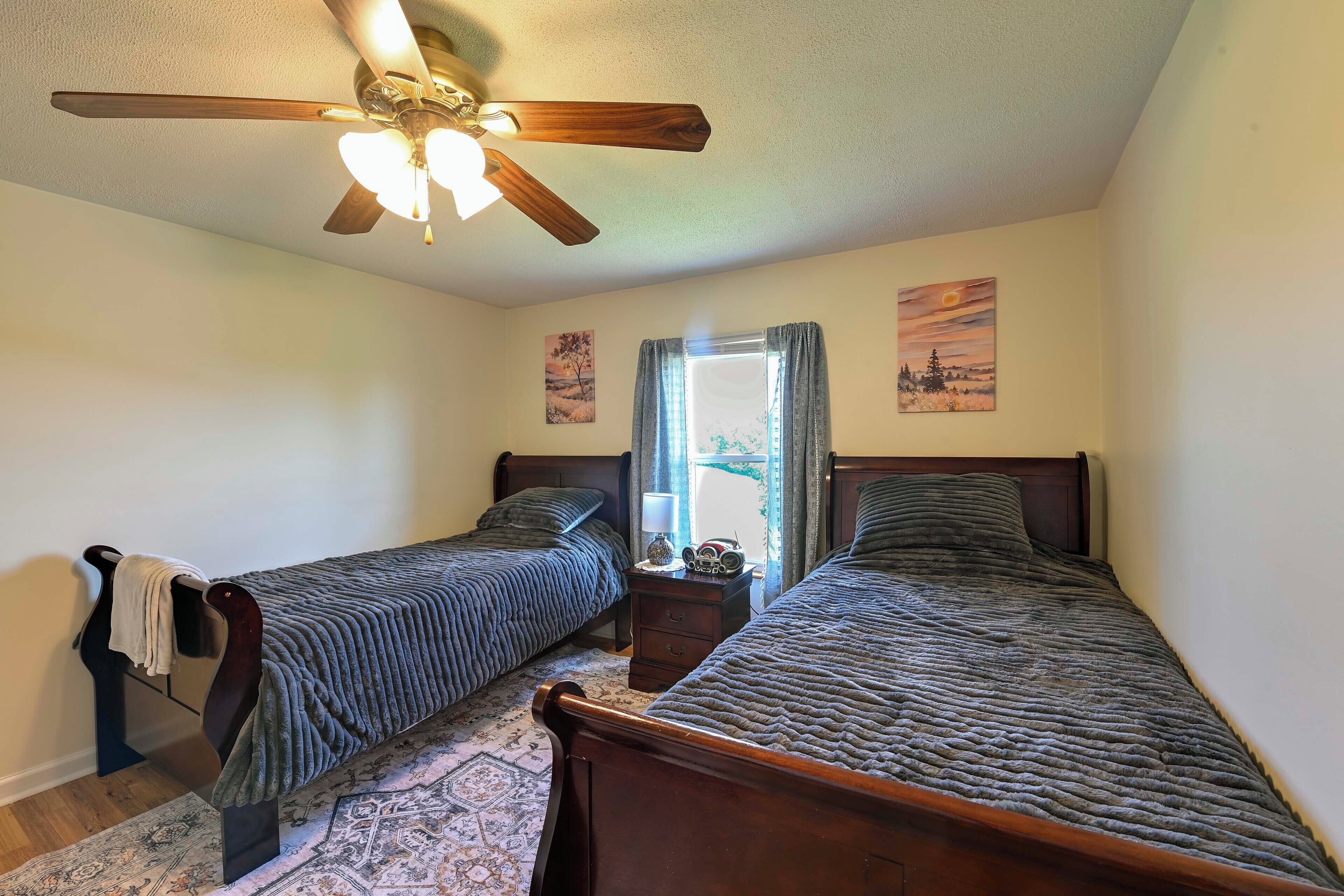 A bedroom with two single beds, a ceiling fan, a window with curtains, and a rug.