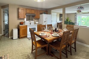 Un comedor con una mesa de madera dispuesta para cuatro personas, rodeada de sillas de madera. Se ven un refrigerador y una estufa blancos al fondo.