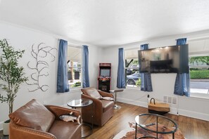 Un salon avec un canapé, un fauteuil, une télévision, une borne d’arcade et une vue sur la rue.