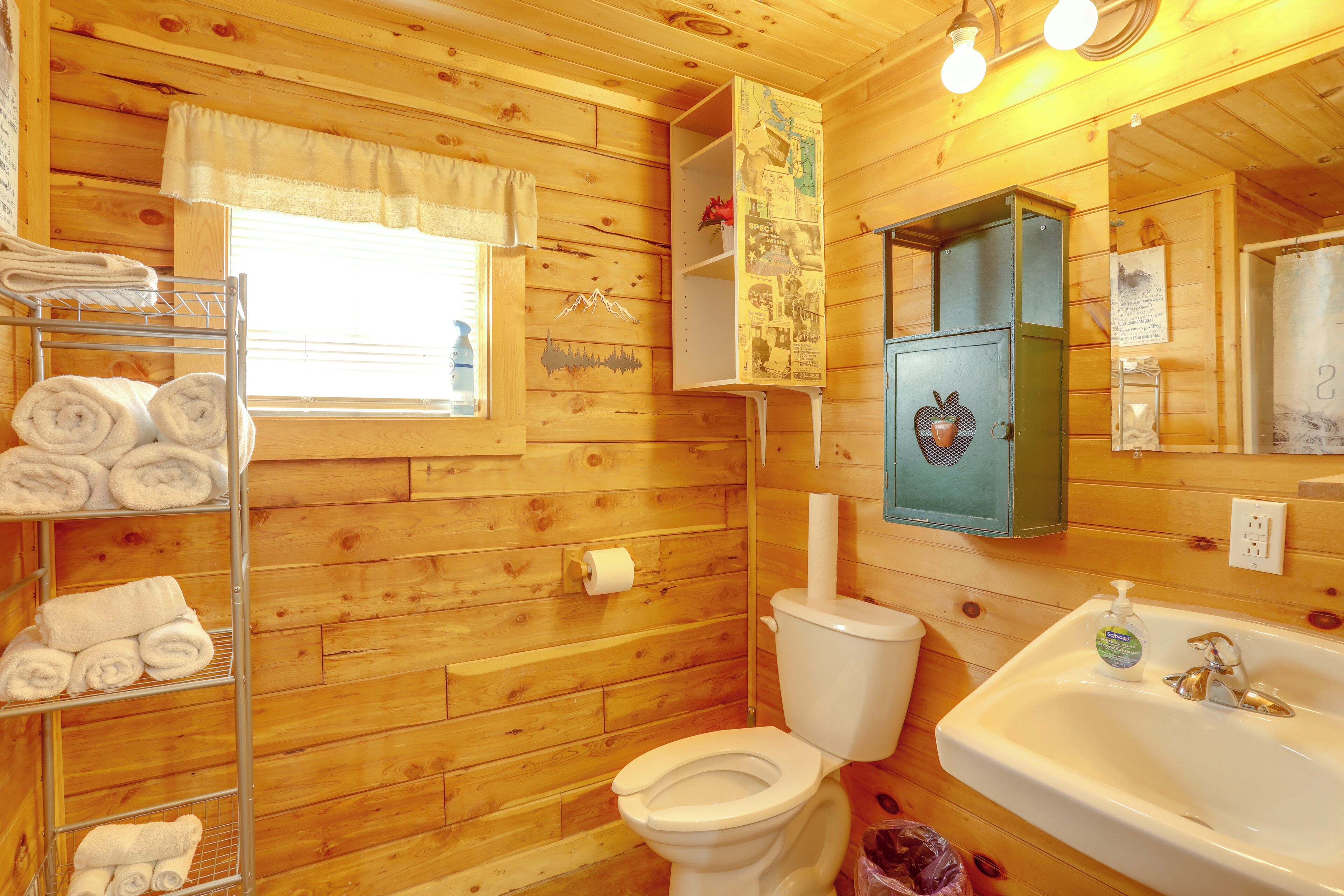 Une salle de bain rustique, aménagée dans une cabane en bois, comprenant des toilettes, un lavabo, un porte-serviettes et une fenêtre avec un rideau.