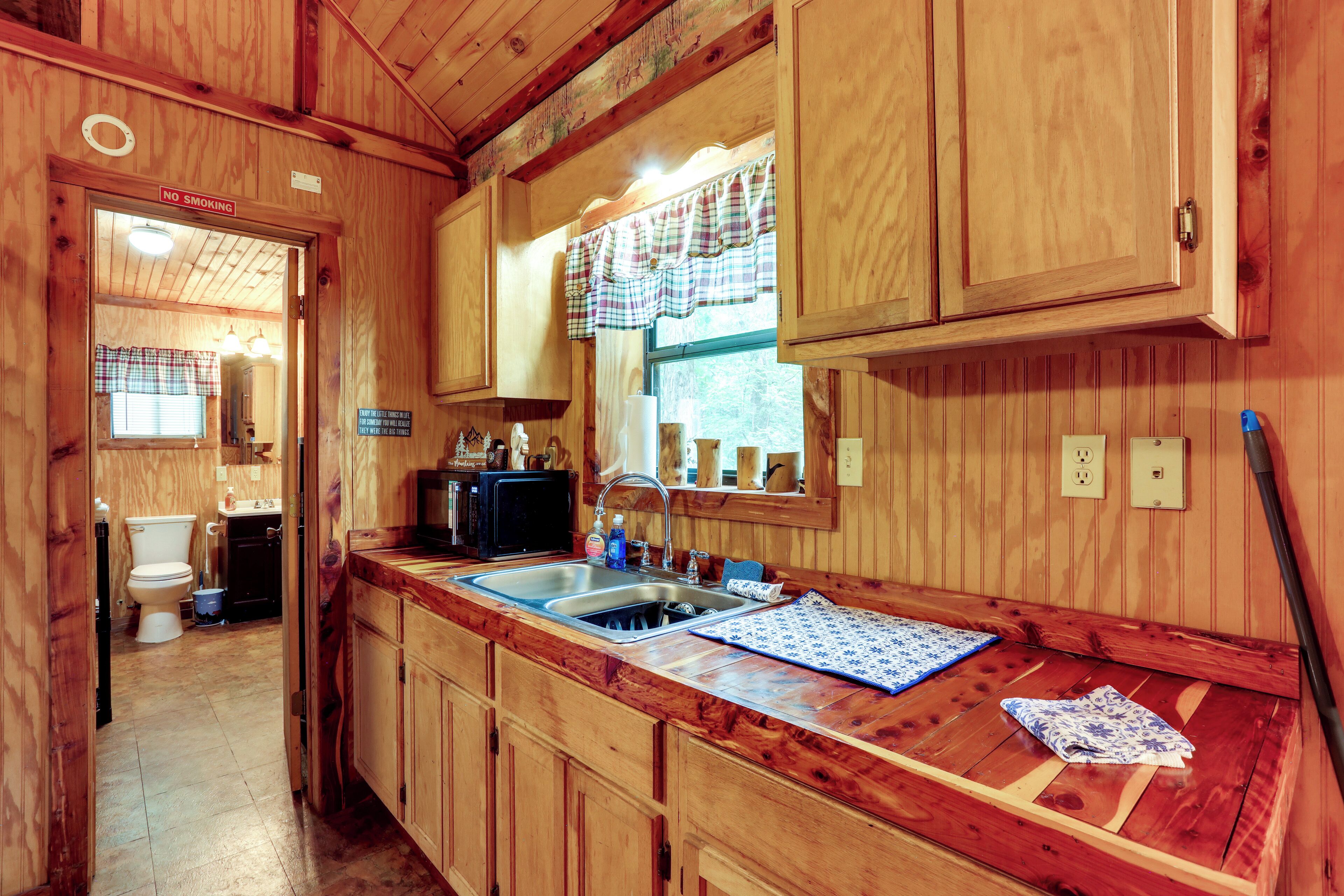 A wooden cabin interior with a kitchen area, including a sink, cabinets, and a window with a plaid curtain.