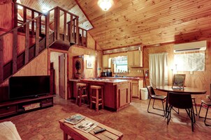 Interior de una cabaña de madera con televisor, taburetes de bar, una mesa de comedor y una zona de cocina.