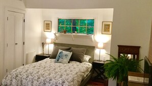 A bedroom with a bed, two bedside lamps, a window with a view of greenery, a door, and a small plant.