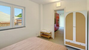 A bedroom with a bed, a small shelf with towels, a wardrobe, and a window with a view of buildings.