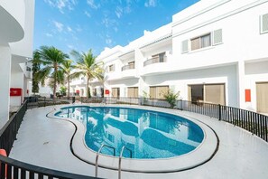 A swimming pool with a curved edge, surrounded by a black fence, and adjacent to a white building with balconies and windows.