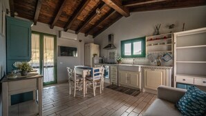 A rustic kitchen with wooden beams, a dining area with white chairs, and a kitchenette with white cabinets. - Atzori Home - Sant'Anna Arresi (Sant`Anna Arresi)