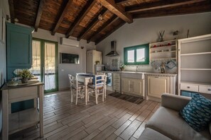 A rustic kitchen with wooden beams, a dining area with white chairs, and a kitchenette with white cabinets. - Atzori Home - Sant'Anna Arresi (Sant`Anna Arresi)