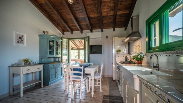A kitchen with wooden beams, a dining area with chairs, and a view through a window. - Atzori Home - Sant'Anna Arresi (Sant`Anna Arresi)