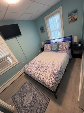 Ein Schlafzimmer mit Bett, Nachttisch, einem an der Wand montierten Fernseher, einem Fenster mit Jalousien und einem kleinen Teppich.