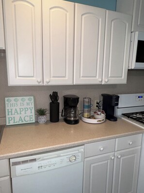 Eine Küche mit weißen Schränken, einer Kaffeemaschine, einer Kaffeekanne, einem Schild mit der Aufschrift 'THIS IS MY HAPPY PLACE' und einem Geschirrspüler.