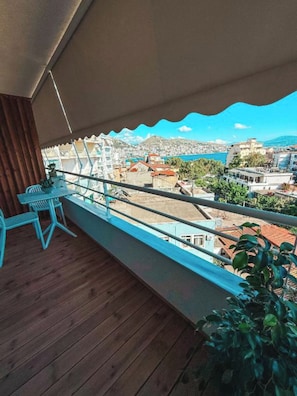 A balcony with a wooden floor, a table, and chairs, overlooking a coastal town with buildings and a body of water. - SIÁMAR Studio Rooms (Saranda)