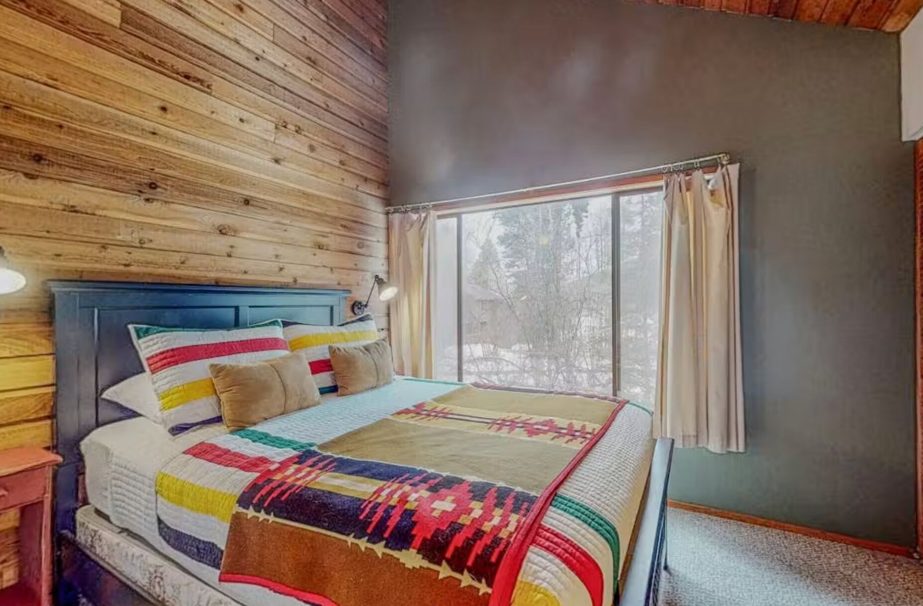 Une chambre avec un mur en bois, un lit avec une literie aux couleurs vives, une table de chevet et une fenêtre avec des rideaux.