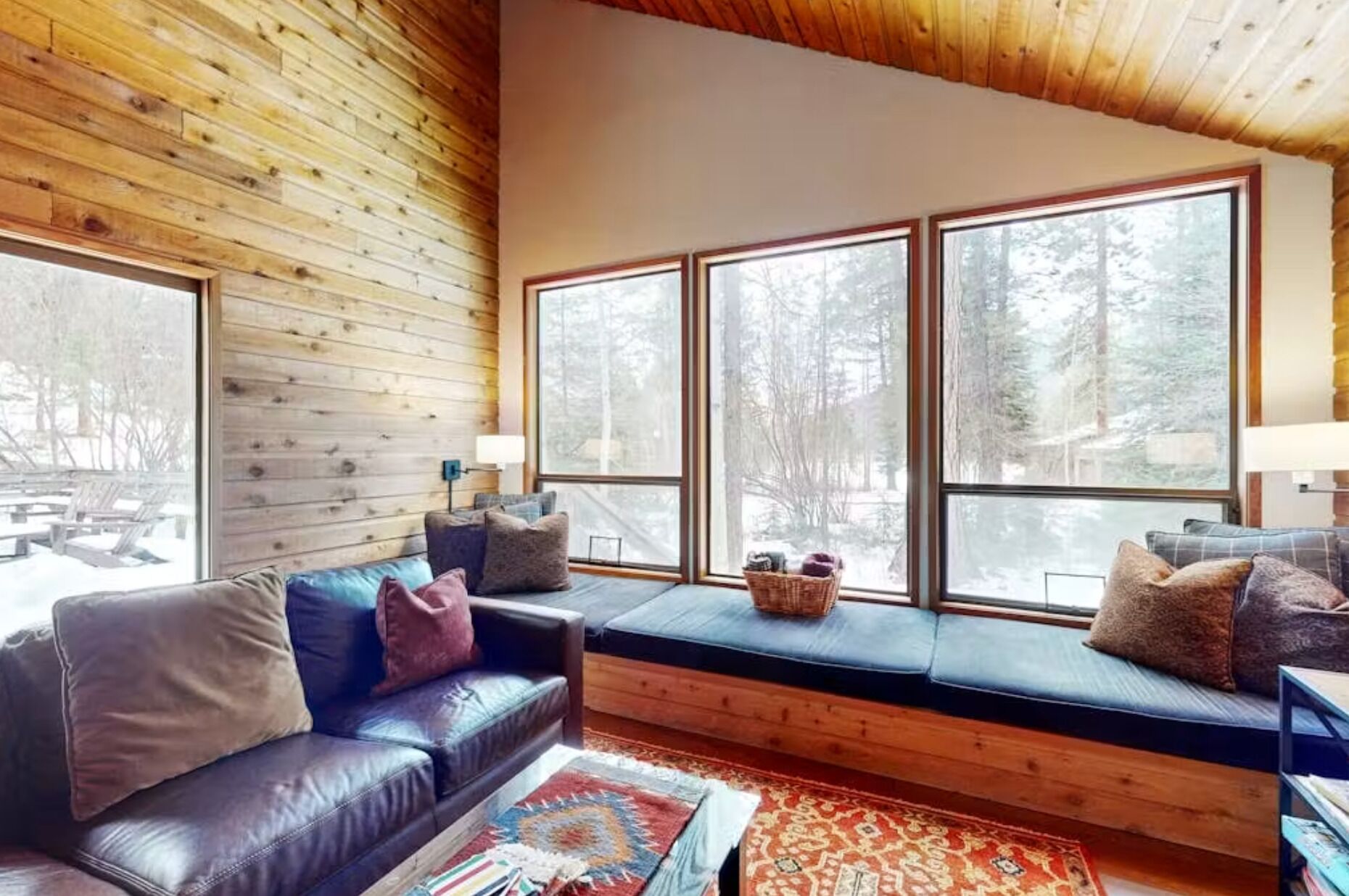 A cozy cabin interior with a wooden ceiling, large windows, a leather sofa, and a bench with cushions.