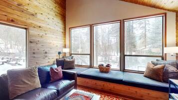 A cozy cabin interior with a wooden ceiling, large windows, a leather sofa, and a bench with cushions.