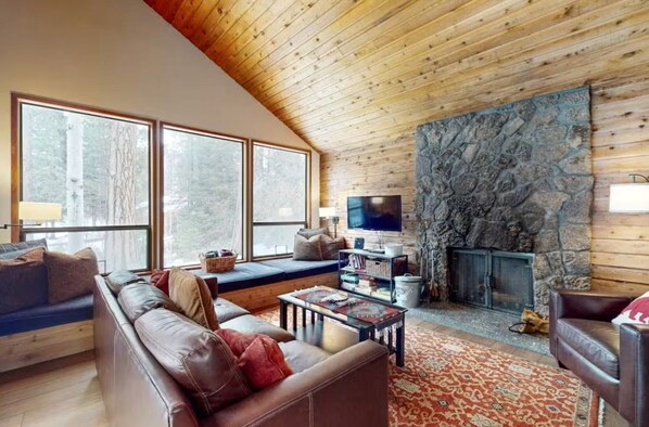 A cozy living room with a stone fireplace, leather sofas, a television, and large windows.