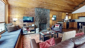 A log cabin interior with a stone fireplace, wooden walls, a sofa, a television, and a kitchen area. - Meadow's Edge at Black Butte Ranch—2 pets allowed (Sisters)