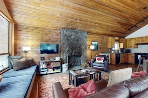 A log cabin interior with a stone fireplace, wooden walls, a sofa, a television, and a kitchen area. - Meadow's Edge at Black Butte Ranch—2 pets allowed (Sisters)
