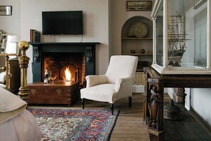 A room with a fireplace, a television mounted above it, a white armchair, a wooden table, and a ship model on a shelf.