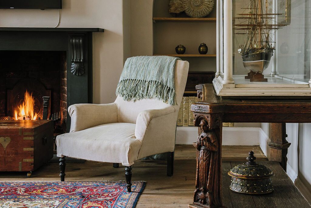A cozy room with a fireplace, a chair, a table with a decorative item, and a ship model on a shelf.