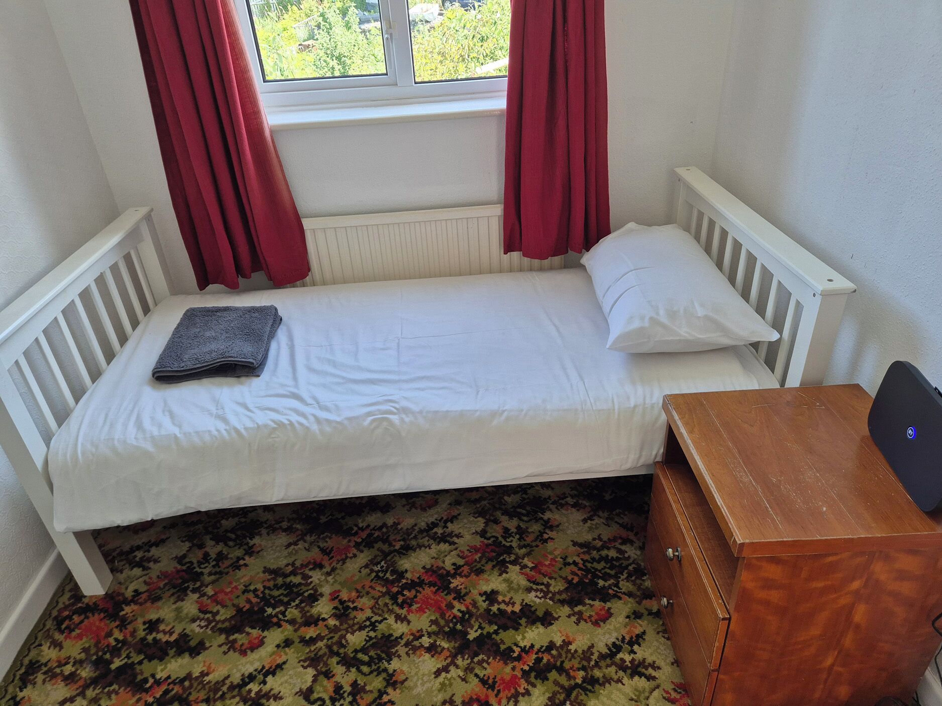 A small, single-bed room with a window, a radiator, a wooden nightstand, and a patterned carpet.