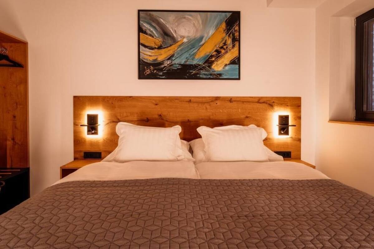 A bed with a geometric patterned bedspread, a wooden headboard, two wall-mounted reading lights, and a painting above the bed.