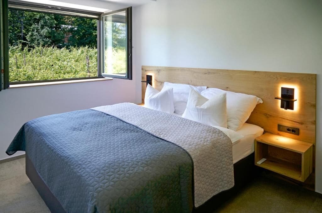Un dormitorio con cama, mesita de noche, luz adosada a la pared y ventana con vistas a zonas verdes.